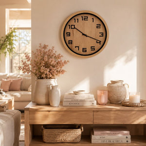 A 40cm Timber Look Wall Clock with a light wood grain face and black numerals, mounted on a beige wall above a wooden console table styled with vases, books, and dried flowers.