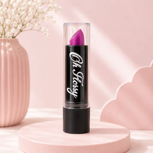 A pink Oh Flossy lipstick bullet in a clear tube with a black base, standing upright on a round pink pedestal. The tube has the words 'Oh, Flossy' in white script.