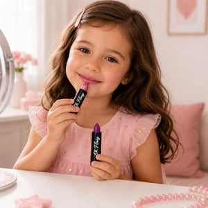 A young girl with wavy brown hair applies Oh FlossyBetter together lipstick while holding another tube of lipstick with a purple tip. She is wearing a pink dress in a bright, soft room with pink accessories on a white table.