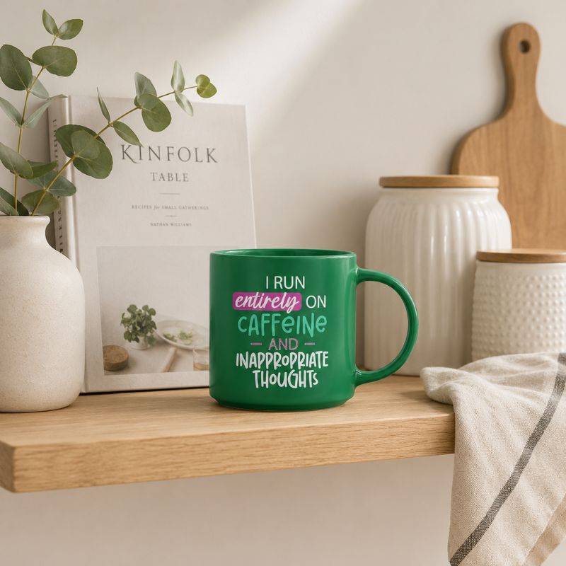 A Cheeky Caffeine Mug with the text 'I RUN entirely ON CAFFEINE AND INAPPROPRIATE THOUGHTS' in white and pink lettering, styled on a wooden shelf with kitchen props like white canisters, a book, and a towel.