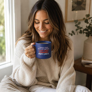 A woman in a cozy beige knit sweater holds a Cheeky Remember Mug with a humorous quote printed on it, set in a bright, sunlit room.
