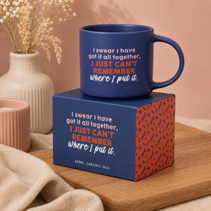 A Cheeky Remember Mug with a humorous quote sits on top of its matching blue and red patterned gift box, styled on a wooden board with a neutral background and dried flowers.