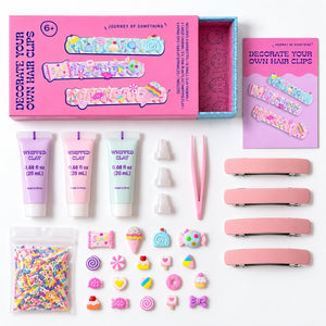 A children's craft kit laid out on a white background, including a pink box labeled 'Journey of Something' with hair clips, three small tubes of whipped clay, tweezers, pink hair clip bars, a bag of multicolor sprinkles, and assorted pastel decorative charms.