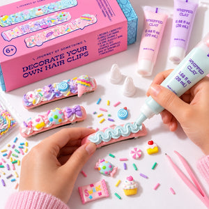 A hands-on craft kit showing decorated pink hair clips with pastel clay decorations, alongside tubes of whipped clay, small candy-shaped accessories, and a pink product box on a white surface.