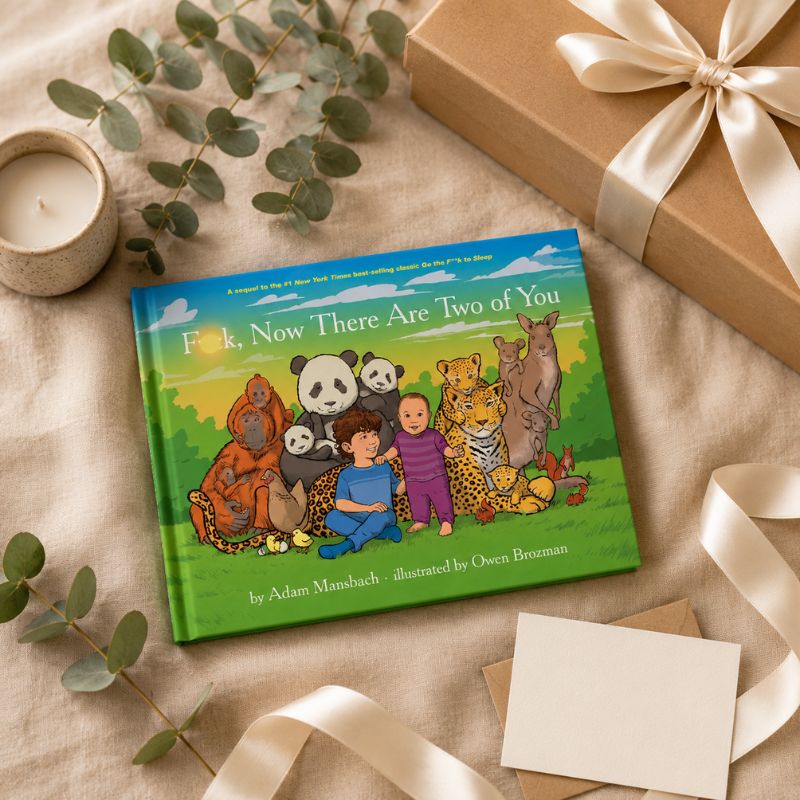 A children's book titled "F*ck, Now There Are Two of You" lies on a beige fabric surface, styled with a wrapped gift box, ribbon, candle, eucalyptus stems, and blank cards. The book cover shows an illustrated group of animals and a child.