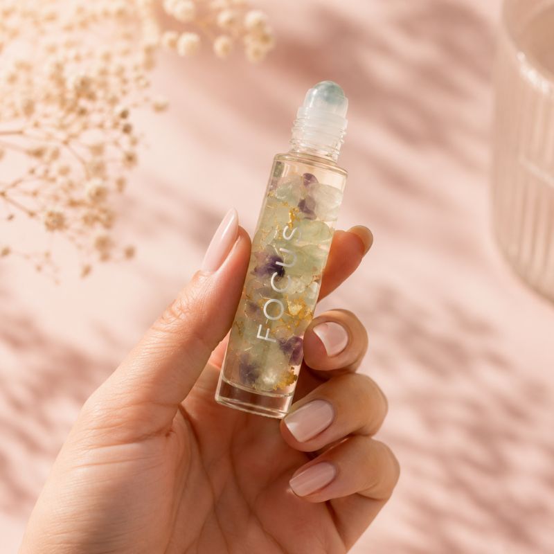 Hand holding a crystal-infused focus rollerball bottle with floral elements against a soft pink background