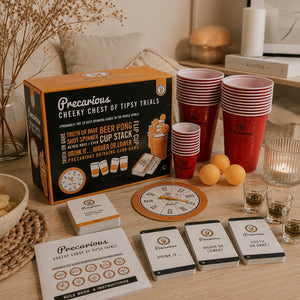 Precarious Cheeky Chest of Tipsy Trials party game set arranged on a wooden table, including the orange and black retail box, red and white cup stacks, drinking cards, a spinner wheel, and small glasses with drinks, in a styled indoor setting.