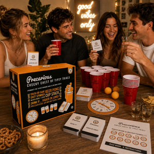 Four people laughing around a table with the Precarious Cheeky Chest of Tipsy Trials game components spread out, including the orange and black retail box, cards, drink tracking board, red cup stacks, and drinks in hand, in a warmly lit indoor setting.