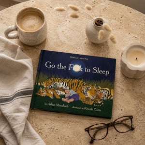 A children's book titled "Go the F*ck to Sleep" by Adam Mansbach, illustrated by Ricardo Corla, placed on a beige stone tabletop with a mug of coffee, a small vase with dried stems, a candle, and eyeglasses around it.