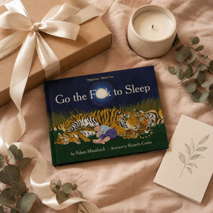 A children's book titled "Go the Fox to Sleep" by Hilarious Mom Fox is styled in a cozy flat lay on a soft beige fabric, surrounded by a wrapped gift with a cream ribbon, a lit candle, eucalyptus leaves, and a small card.