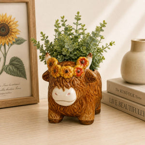 Ceramic cow planter with greenery on a wooden surface with books and a sunflower painting in the background.