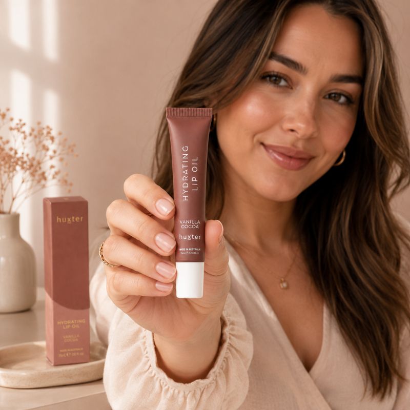 Woman holding Huxter Vanilla Cocoa Hydrating Lip Oil in warm neutral lifestyle setting