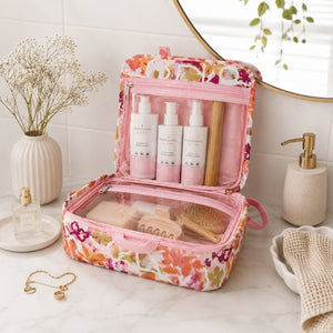A Large hanging toiletry bag in Ivy floral print with pink and orange details is shown open on a bathroom counter, filled with skincare bottles, a brush, and other beauty items. The scene includes a vase of dried flowers, a perfume bottle, a soap dispenser, a towel, and a mirror in a bright, clean bathroom setting.