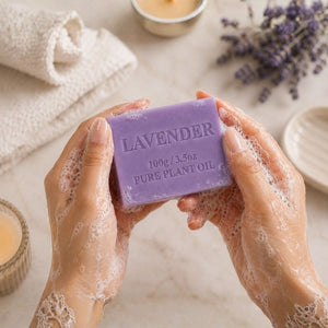 Hands holding a lavender soap bar with lavender flowers and candles in the background