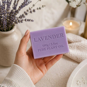 Hand holding a Lavender Soap Bar 100g with a blurred background of lavender flowers and a candle.