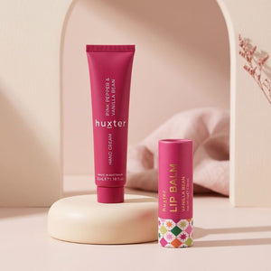 Huxter Pink Pepper and Vanilla Bean hand cream with Vanilla Bean lip balm on a warm blush studio pedestal