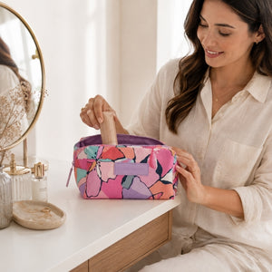 A woman in a white linen shirt sits at a vanity table, placing a makeup sponge into a Makeup Cosmetic Bag – Willow. The pouch features a vibrant pink, purple, and orange abstract floral pattern with a purple zipper and a lavender brand tab.