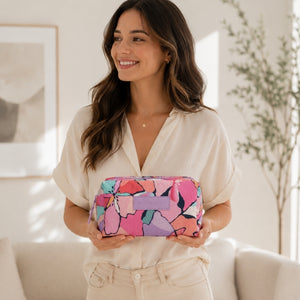 A woman in a cream blouse holds a MakeUpp Cosmetic Bag - Willow with a pink, purple, and blue abstract floral pattern in a bright, modern living room setting.
