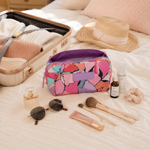 A MakeUp Cosmetic Bag - Willow with a purple interior, styled on a cream bed alongside an open suitcase, a straw hat, sunglasses, makeup brushes, a perfume bottle, and other travel accessories.