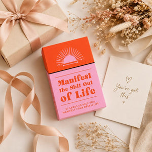A pink and orange card deck box labeled 'Manifest the Shit Out of Life' is styled on a light beige surface with a kraft gift box, ribbon, dried flowers, and a cream card that reads 'You've got this'.