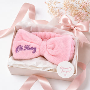 A pink plush bow headband with purple embroidered text reading 'Oh Flossy' inside a light pink gift box with tissue paper, pink satin ribbons, dried flowers, and a round sticker that says 'Especially for you'.