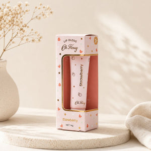 A pink lip gloss tube in a matching pink and gold box, labeled 'Oh Flossy' and 'Strawberry', displayed on a round stone tray with dried flowers and soft sunlight in the background.