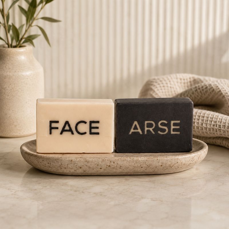 Two soap bars labeled 'FACE' and 'ARSE' on a stone tray with a plant and towel in the background.