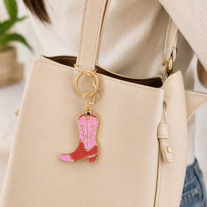 A close-up of a beige tote bag worn by a person, featuring a Pink Cowboy Boot Enamel Keyring with pink and red enamel and gold-tone metal.
