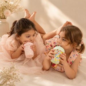 Two young girls sitting on a bed, each holding a Pink Daisy Mini Smoothie Cup & Straw 200ml