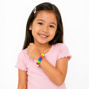 A young girl with long dark brown hair smiling, wearing a pink short-sleeve t-shirt and a Pom Pom Squishy Bracelet on her wrist, is set against a plain white background.