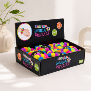 A black retail display box filled with colorful Pom Pom Squishy Bracelet, photographed on a white tabletop with a vase and chair in the background. The box is labeled 'Pom Pom SQUISHY BRACELET' and shows the brand 'mii'.