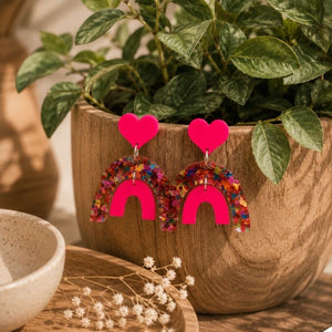 Pop Rainbow Dangle Earrings with pink arches on a wooden surface with a plant in the background.
