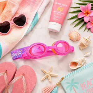Rock Candy Pink Swim Goggles styled with summer beach essentials including sunscreen, sunglasses, tropical flowers, and a pastel towel in a bright flat lay composition.