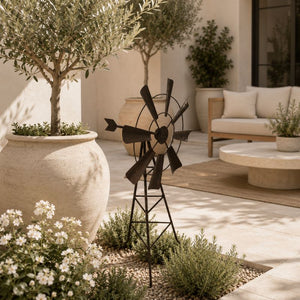 Metal garden windmill stake displayed on a stylish outdoor patio with beige seating, potted olive trees, and white flowers.