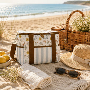 A Saddle & Bloom cooler bag sits on a sandy beach picnic setup, surrounded by a straw hat, sunglasses, a rolled cream towel, lemons, a wicker basket with flowers, and the ocean in the background.