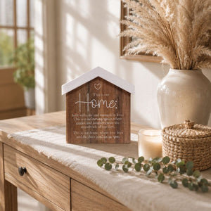 A Saddle & Bloom Home Blockword is displayed on a rustic wooden table in a warm, sunlit room. The scene includes a beige ceramic vase with dried pampas grass, a lit candle, and a woven basket.