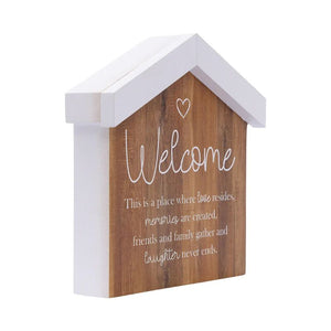 A house-shaped wooden wall decor sign with a white painted frame and a natural wood front panel. The panel features the word 'Welcome' in white script, a small heart outline, and a sentimental quote below, all set against a plain white background.