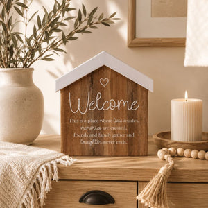 A Saddle & Bloom Welcome Blockword 'Welcome' lettering sits on a wooden dresser in a warm, styled room. The sign includes a heart and a quote about home.