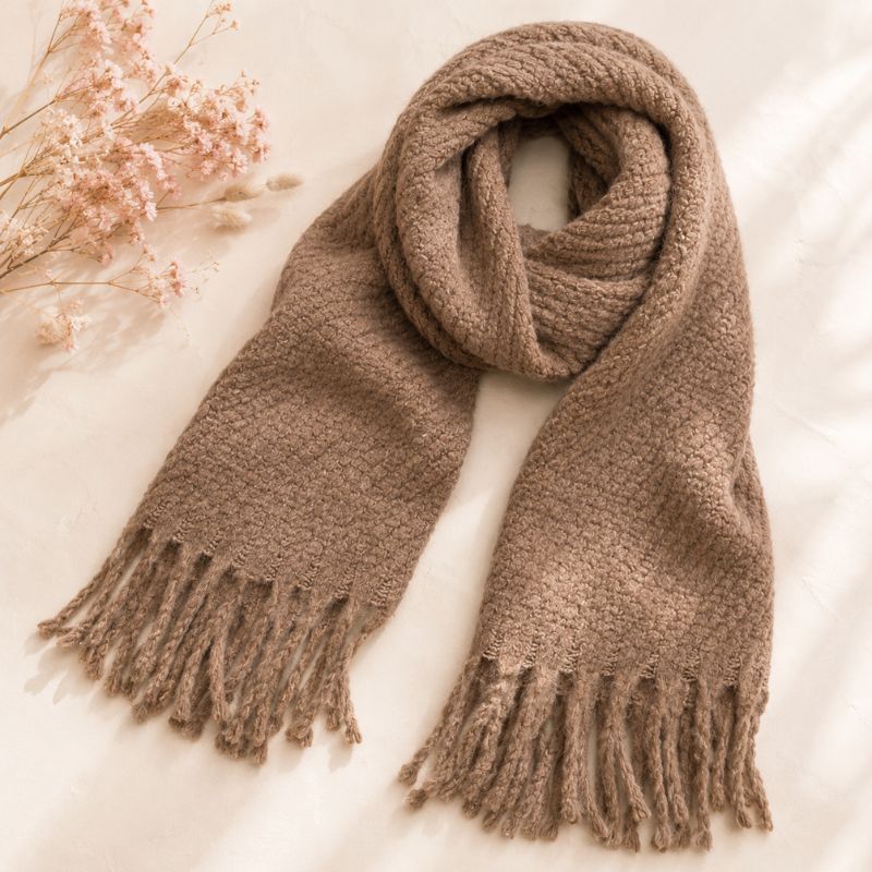 A Sand Chunky Knit Winter Scarf with Fringe, styled in a loop with fringed ends on a soft cream fabric background with dried flowers in the corner.