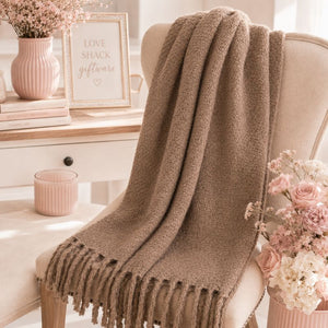 A Sand Chunky Knit Winter Scarf with Fringe is draped over a cream upholstered armchair in a bright, styled room. A white side table, pink ribbed vase with flowers, and a framed sign reading 'LOVE SHACK giftware' are visible in the background.