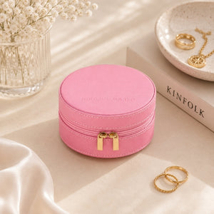 A Small Jewellery Case – Berry with a gold zipper and dual pull tabs, styled on a beige surface with decorative props including rings, a vase of flowers, and a book.