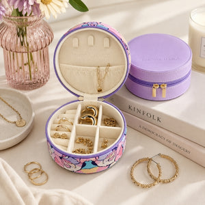 An open round jewelry box with a floral print exterior and cream interior organizer, shown with gold rings and earrings on a draped fabric surface. A closed matching jewelry case sits beside it, along with decorative books, a pink glass vase, and scattered jewelry.