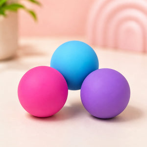 Three matte silicone massage balls in pink, blue, and purple, arranged together on a light beige surface with a soft pink background.