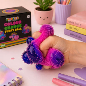 Smoosho’s Colour Change Furry Ball being squeezed in a hand on a pastel desk, with the product box, plant, books, and stationery in the background.