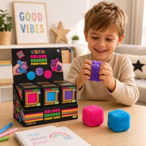 A young boy smiling and squeezing a Smoosho's Colour Change Furry Cube on a wooden table, with two other squishy cubes in pink and blue beside him and a display box of GooGoo Colour Change Furry Cube toys behind him.