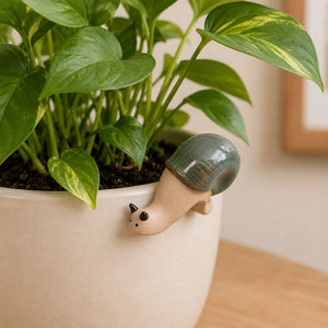 A small ceramic snail garden stake with a glossy green shell and beige body, growing out of the side of a light beige planter filled with green trailing houseplant leaves.-Snail Pot Hanger Green 8cm