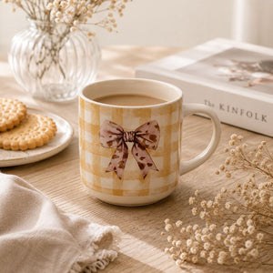 A Splosh -Saddle & Bloom Bow Mug. The mug is styled with dried flowers, cookies on a plate, a glass vase, and a book in a warm, cozy indoor setting.