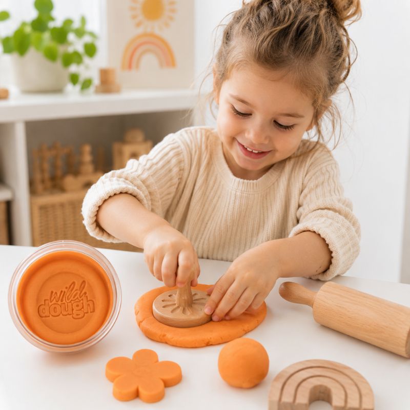 Smiling child playing with Wild Dough orange sensory playdough using a wooden rolling pin and play tools.
