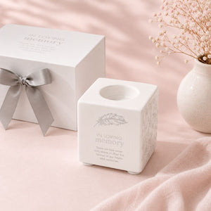 White ceramic candle holder with 'In Loving Memory' text on a pink surface with a gift box and vase.