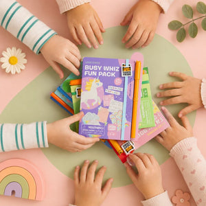 Children's hands reaching towards a Busy Whiz Fun Pack on a pink and green table.